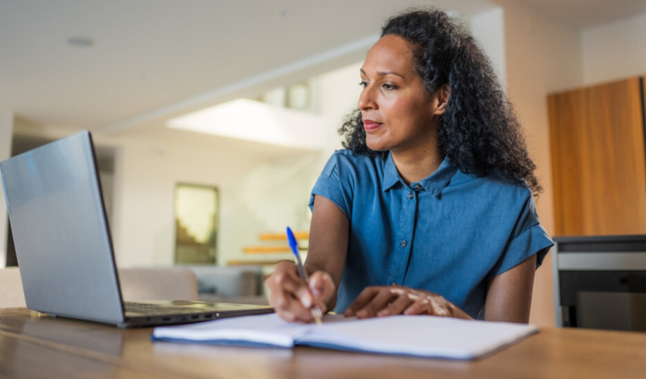 Adult women at a desk with a laptop, writing in a notebook