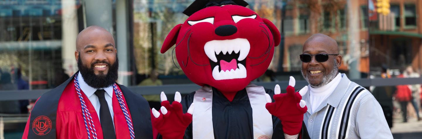A male student in his cap & gown, Pounce and a dad at the 2025 DU Commencement ceremony