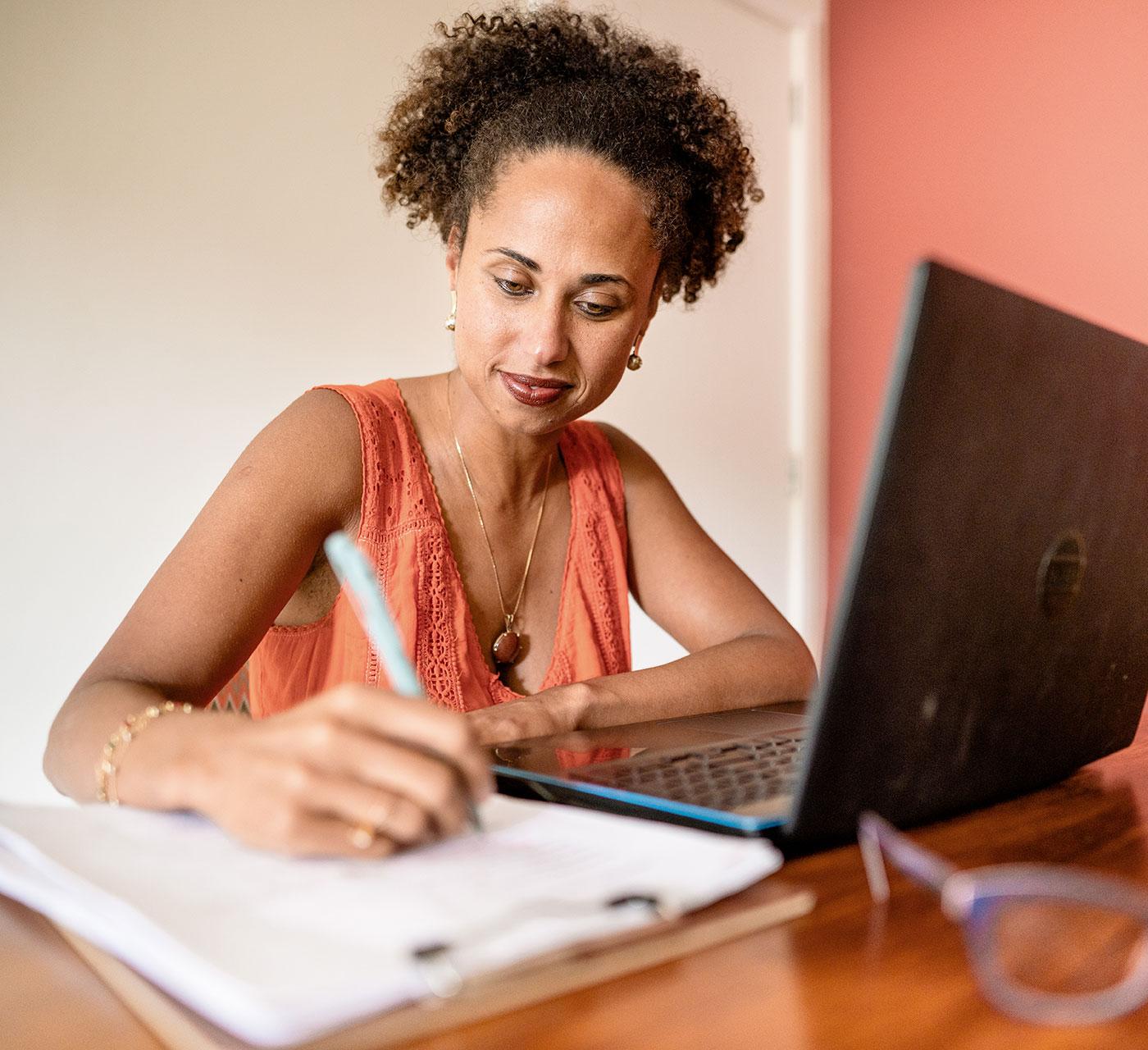 Adult woman with curly hair using laptop and writing notes