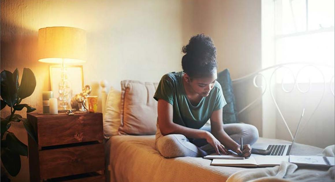 A student sitting on a bed with a laptop and notebook